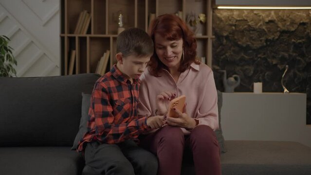 Mother learning her autistic son how to use mobile phone sitting on the couch at home. Boy with autism using smartphone with mom's help.