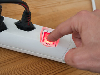 Pressing the red illuminated button of a white power strip, on a wooden background.