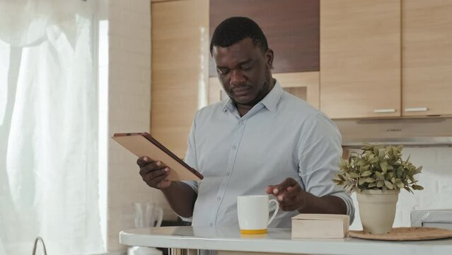 Medium shot of Black man in light blue office shirt reading news on digital tablet while drinking his morning coffee at kitchen, getting ready for work