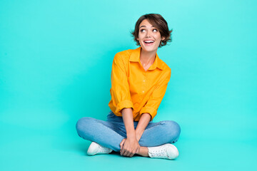 Full length photo of cheerful lady sit floor leg crossed interested look empty space offer promo banner isolated on cyan color background