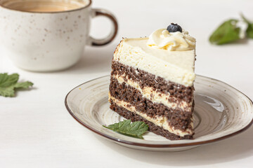 A piece of layered cake with white cream on the plate, a white ceramic cup, green leaves on white table, delicious dessert concept
