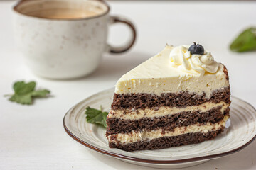 A piece of layered cake with white cream on the plate, a white ceramic cup, green leaves on white table, delicious dessert concept