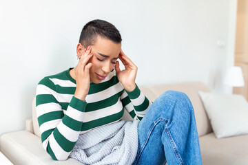 Portrait of an attractive woman sitting on a sofa at home with a headache, feeling pain and with an expression of being unwell. Upset depressed woman lying on couch feeling strong headache migraine.