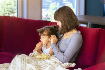 Mom and daughter are sitting on a bright sofa and eating popcorn together. They are covered with a warm blanket. The concept of comfort, love and mutual understanding in the family