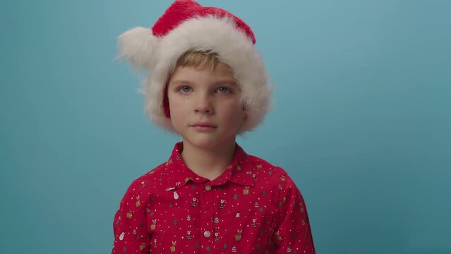 Portrait of frustrated kid in Santa's hat crying looking at camera standing on blue background. Child upset with Christmas.