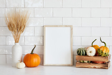 Picture frame mockup with wooden box of decorative pumpkins, vase of dry wheat on white table in...