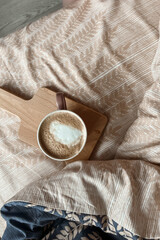 On the bed with beige bedding there is a wooden board with a pink mug of coffee with milk foam. Top view