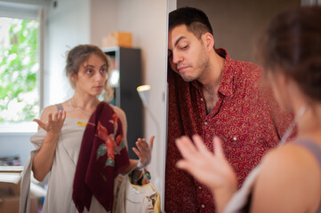 Charming young woman is trying on clothes and having fun standing in front of the mirror in the bedroom, but the man is exhausted and bored