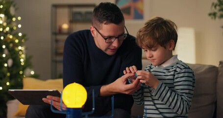 Son is spending time with dad men are sitting on couch playing with a model of planetary system guy is explaining to boy about construction of cosmos using toy as example he is naming planets.