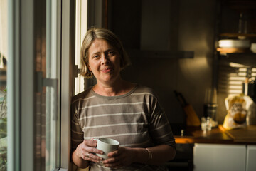 mid adult woman standing by the kitchen window and drinking coffee