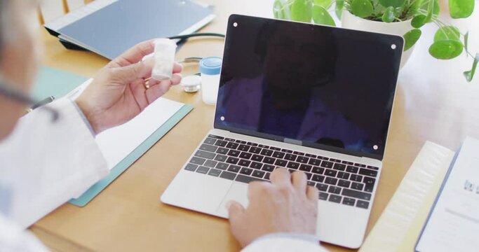 Senior biracial male doctor using laptop and holding medication, copy space on screen