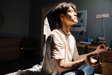 Young asian man wearing t-shirt meditating while sitting on bed