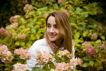 Naklejka premium beautiful smiling brunette girl in pink sport jacket sitting in autumn park in bushes of pink hydrangeas