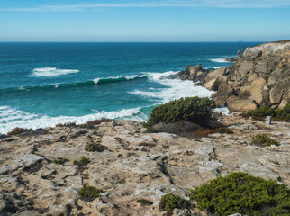 View sea shore with ocean waves, sharp rocks and stones and green bush at Rota Vicentina wild coast near Vila Nova de Milfontes, Portugal.