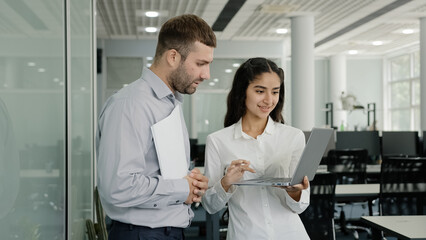 Positive friendly colleagues chatting in office discussing project details young female employee consults with male mentor shows presentation on laptop girl manager communicates co-worker in workplace - obrazy, fototapety, plakaty