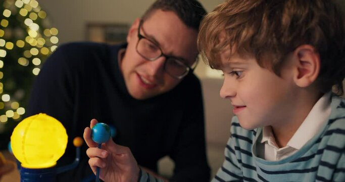 Close-up of seven-year-old boy and dad, who are considering model of the solar system. The boy is holding model of the planet Earth, and father with glasses tells him about it. Boy listens intently.