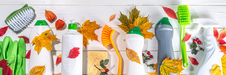 Autumn cleaning flatlay, advertising banner mockup for autumn cleaning campaign. Set of various detergents, bottles, spray, accessories, utensils, white wooden background with autumn leaves and decor