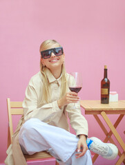 Stylish blonde girl in beige coat and sunglasses sits at wooden table with glass of red wine in her hands on pink background