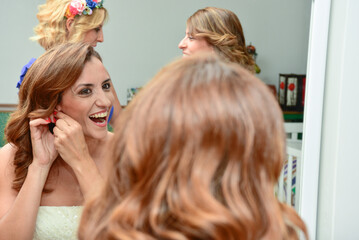 Fototapeta premium Bride looking in the mirror and putting on her earrings. Concept of getting ready in the morning for the wedding day.