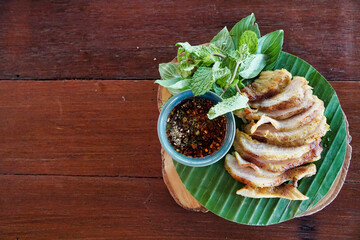 Thai food called Thai Grilled Pork Neck with Spicy Dipping Sauce is in a plate placed on a wooden table placed on the right side of the picture.