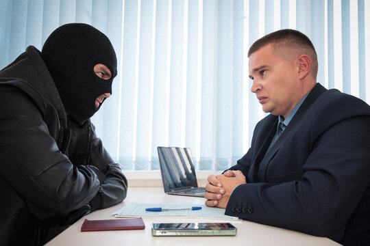 businessman and robbers are sitting at a table. A racketeer in a black balaclava forces to sign a contract. The concept of a raider takeover of the company. signing of important documents.