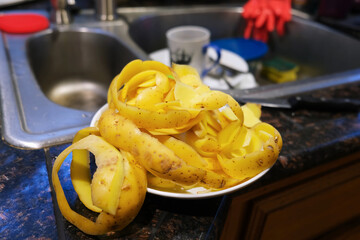 Raw potato peel heap in plate on kitchen table at sink with dirty dish close up photo. Food waste. Home chores, cooking, cleaning, dishwashing concept. Kitchen interior and equipment.