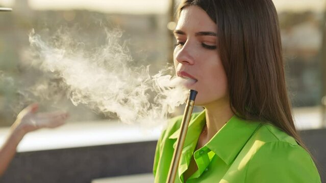 Brunette girl in green shirt smoking hookah. Beautiful lady breathing out thick white smoke. Close up. Sunny blurred backdrop.