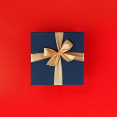 Blue gift box with light golden ribbon on festive red background. Xmas and New Year postcard design. Black Friday sales, Birthday celebration party concept