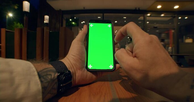 Close up view of young male tattooed hands with watches holding vertical and using mobile phone with green screen, scrolling smartphone touchscreen on table with soft lights of cafe on background