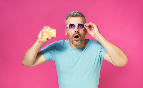 Excited of a new offer handsome man hold debit, credit card in hand lowering his blue glasses wearing blue t-shirt isolated on pink background. Man with bank card in hand. Financial, banking concept