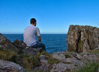 Naklejka na ściany i meble Young man sitting on the edge of a cliff thinking and looking at the sea