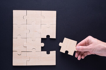 Hand attaches the missing piece of the puzzle. Business concept with wooden jigsaw puzzle on black...