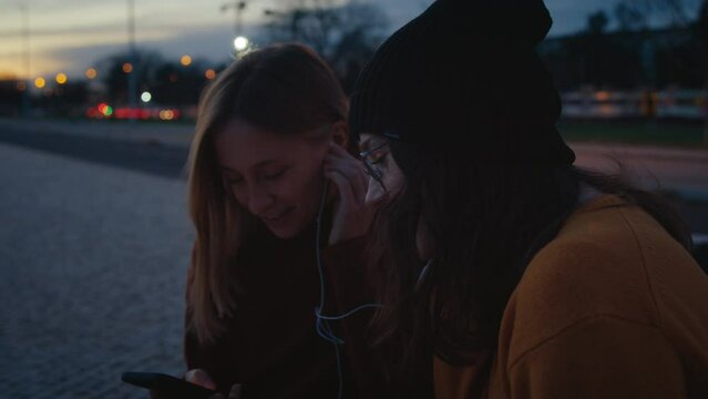 Two best female friends hang out at sunset in park, listen to music and dance to beats. Music streaming platform or application, two women share headphones and enjoy, smile and laugh 