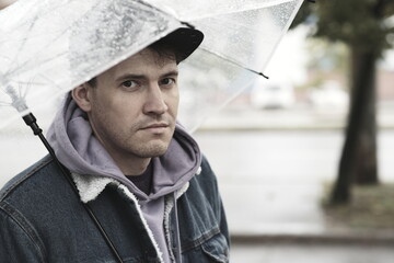 Young pensive man standing with transparent umbrella in rain on city street.