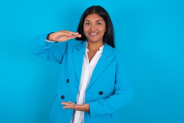 Young latin woman wearing  blue blazer blue background gesturing with hands showing big and large size sign, measure symbol. Smiling looking at the camera.