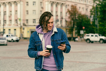 Fototapeta premium Hipster man with dreadlocks drinking hot coffee on street together with cell phone Outdoors city background. Young male in warm clothes drinking hot beverage