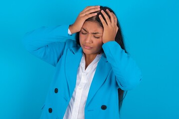 Young latin woman wearing  blue blazer blue background suffering from strong headache desperate and stressed because of overwork. Depression and pain concept.