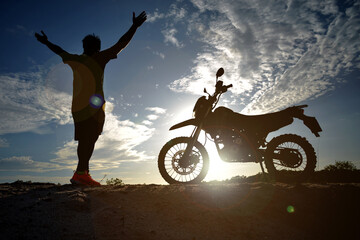 A man raising his hands happily and freely with a motocross bike next to it.
