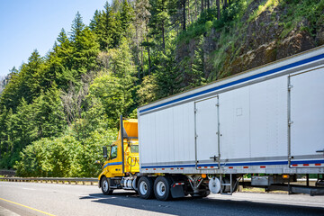 Day cab yellow big rig semi truck with roof spoiler driving with dry van semi trailer on the spectacular highway road with forest on the mountain