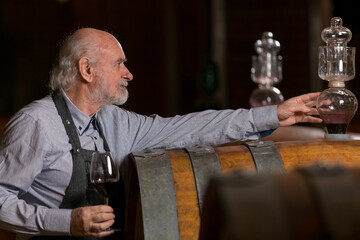 Senior man winemaker at winery checking barrels in wine cellar.