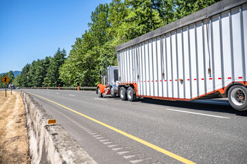 Orange and white big rig day cab semi truck transporting cargo in covered bulk semi trailer with stiffening rib
