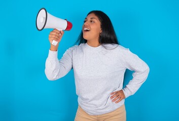 Funny Young latin woman wearing gray sweater blue background People sincere emotions lifestyle concept. Mock up copy space. Screaming in megaphone.