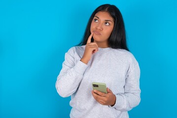 Young latin woman wearing gray sweater blue background thinks deeply about something, uses modern...