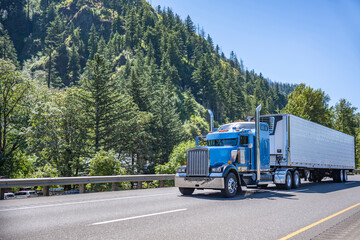 Powerful blue big rig long haul semi truck transporting frozen cargo in reefer semi trailer running on the wide highway road in Columbia Gorge area