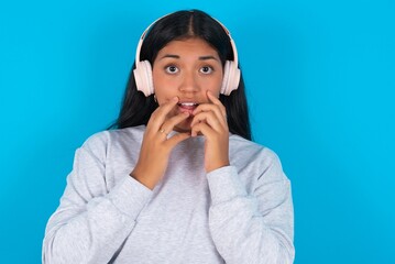 Shocked Young latin woman wearing gray sweater blue background stares fearful at camera keeps mouth widely opened wears wireless stereo headphones on ears