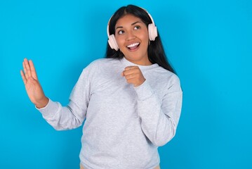 Happy Young latin woman wearing gray sweater blue background sings favourite song keeps hand near mouth as if microphone wears wireless headphones, listens music