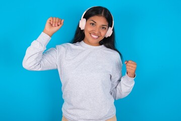 Carefree Young latin woman wearing gray sweater blue background with toothy smile raises arms dances carefree moves with rhythm of music listens music from playlist via headphones