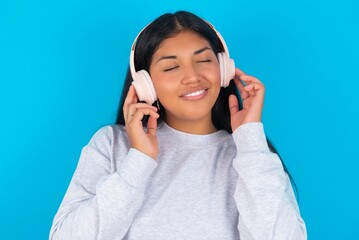 Young latin woman wearing gray sweater blue background with headphones on her head, listens to music, enjoying favourite song with closed eyes, holding hands on headset.
