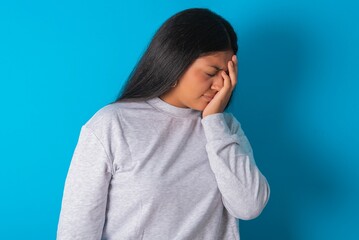 Young hispanic woman wearing grey sweatshirt over blue background with sad expression covering face...