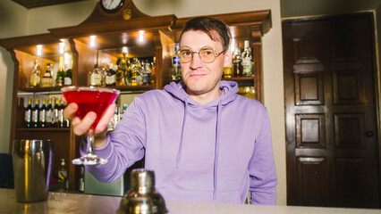 Male bartender in eyeglasses with cocktail standing behind bar, looking at camera, offering alcoholic beverage, smiling.